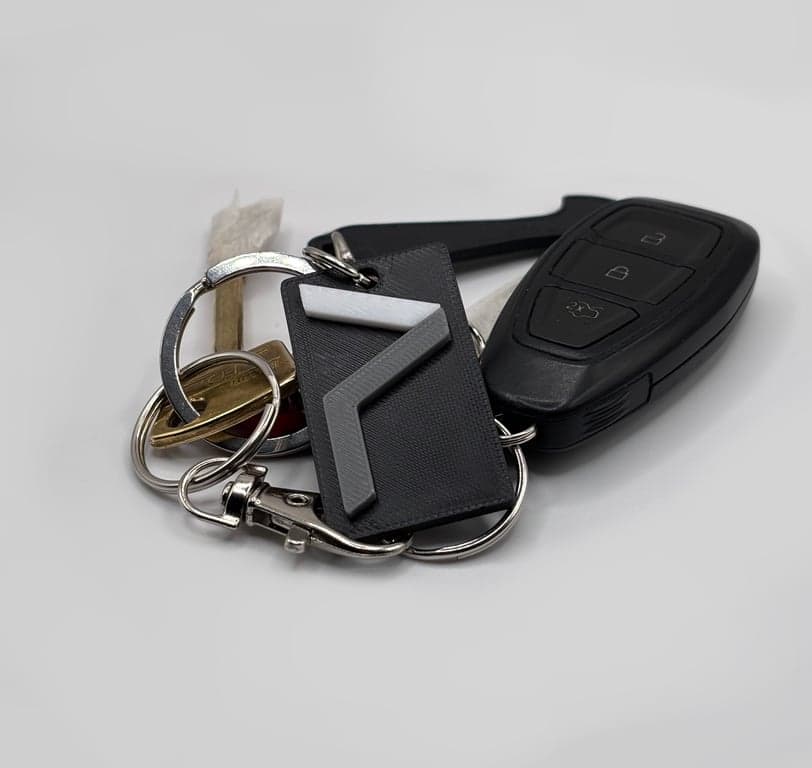 Close-up of a keychain with multiple keys and a black car key fob. The keychain includes a metallic key ring, a brass key, and is focused on a black rectangular keychain accessory with a geometric Printeresting Logo.